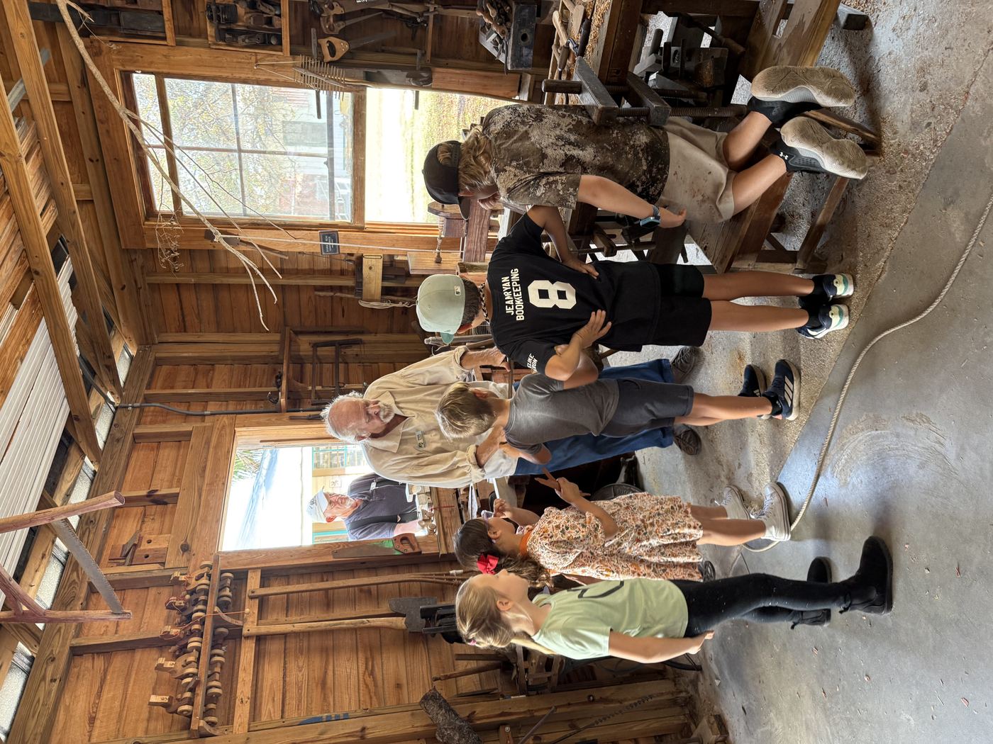 Group of homeschool students gathered around a craftsman demonstrating traditional woodworking at a historic site